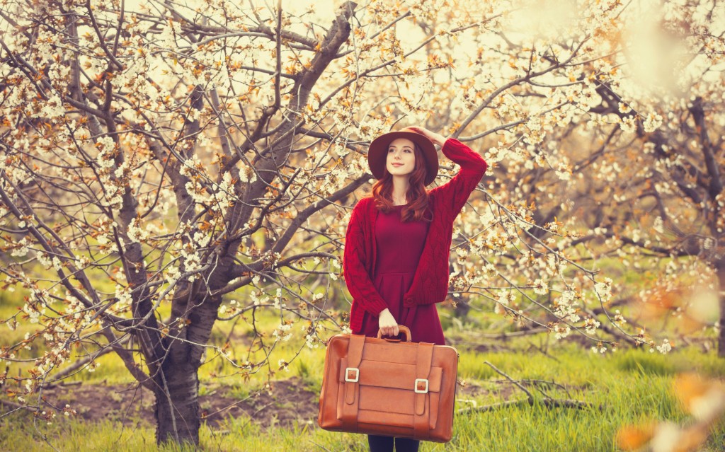 Portrait of a beautiful redhead women in red sweater and hat with suitcase in blossom apple tree garden in spring time on sunset.