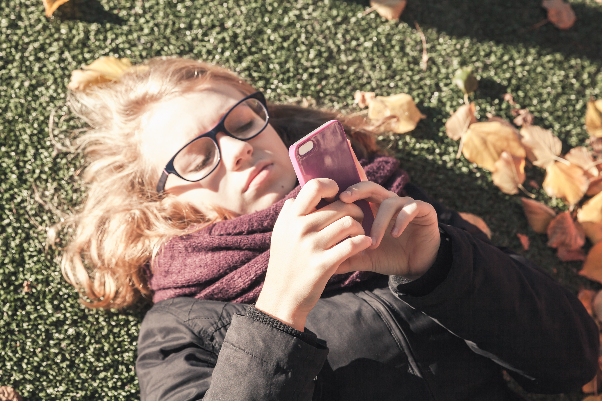 Caucasian blond teenage girl laying in autumnal park and using smart phone, vintage style tonal correction photo filter