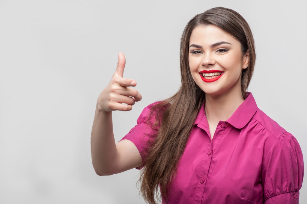 Waist up portrait of beautiful girl reporter with Caucasian appearance, who is smiling and looking at the camera and raising her right hand in such way as if she were trying to shoot someone in jest, isolated on a grey background and there is copy place in the left side
