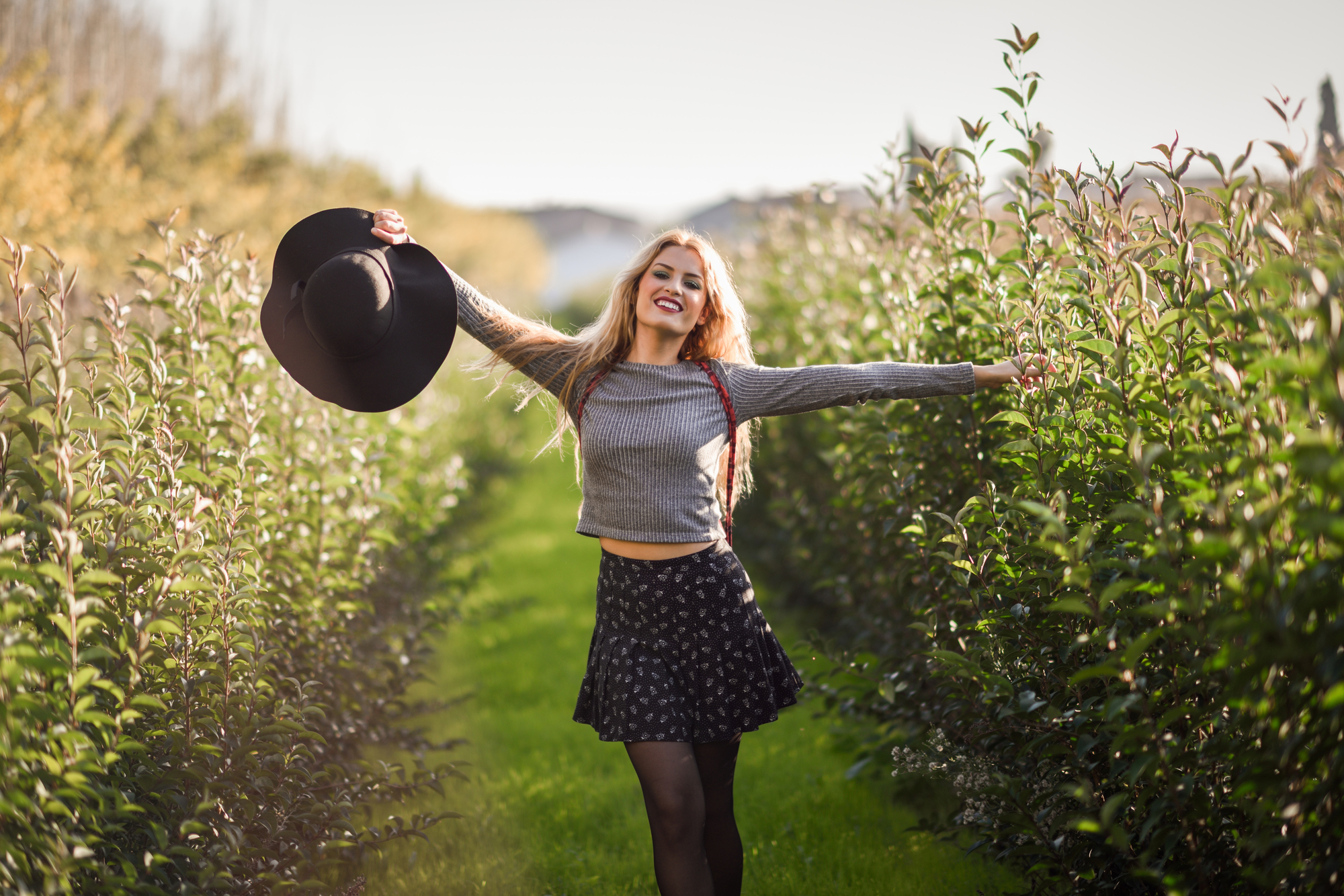 Portrait of blonde young woman happy with open arms in rural road. Girl wearing swater, skirt and hat