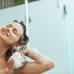 Happy woman washing hair with shampoo under water jet